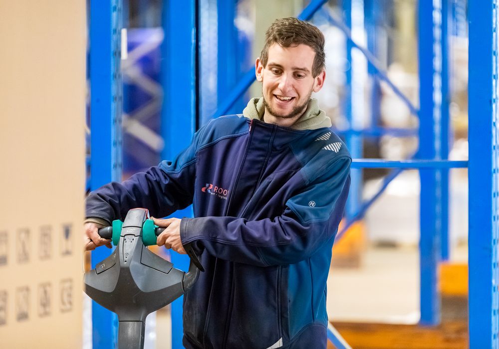 Lachende jonge magazijnmedewerker van De Rooy Transport met een elektrische palletwagen